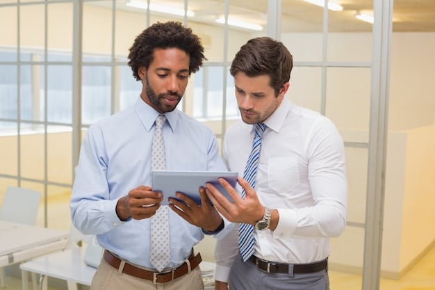 Two colleagues in an office setting, one explaining a task to another, and both looking at a tablet. The image illustrates delegation and collaborative problem-solving.