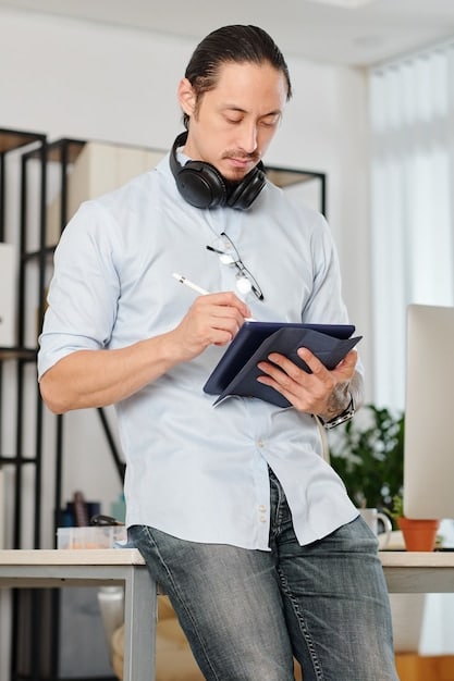 A person wearing headphones using a stylus on a tablet, with a task management app featuring colorful project boards and timelines displayed on the screen. There is an organized workspace with sketchbooks and art supplies scattered around, blending digital with analog tools.