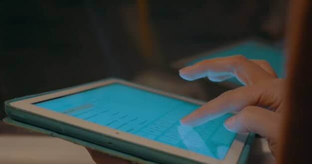 A close-up shot of a person's hand using a stylus on a tablet, which displays a digital Eisenhower Matrix with color-coded tasks. The background is slightly blurred, suggesting a productive workspace.